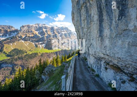 Description: Male tourist walks on alpine trail from Äscher to Ebenalp under a big rock wall with Hoher kasten mountin in the background. Seealpsee, A Stock Photo