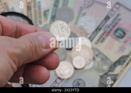 Cropped hand of a man holding a coin against a background of various coins and paper dollar bills. Stock Photo
