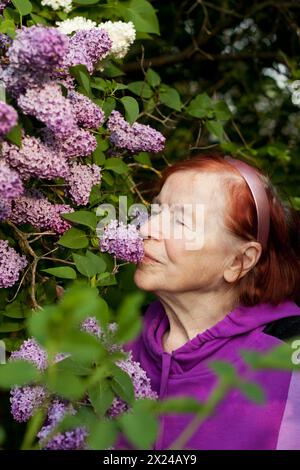 Outdoor close-up portrait of old woman. Beautiful elderly woman against background of blooming lilacs in spring park. Elderly lady woman enjoying scen Stock Photo