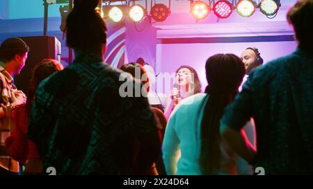 Happy woman doing karaoke at party, singing songs with DJ in front of friends on dance floor. Confident girl having fun with live show perfromance next to electronic mixing station at nightclub. Stock Photo