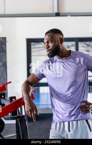 African American young male athlete leaning on gym equipment in gym, taking a break. He has short black hair, a beard, and is wearing a purple shirt. Stock Photo