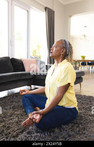 Senior African American woman, grey braids, in yellow shirt, meditates at home Stock Photo