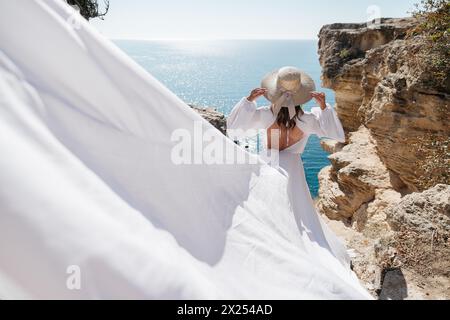 A woman in a white dress is standing on a rocky cliff overlooking the ocean. She is wearing a straw hat and she is enjoying the view. Stock Photo