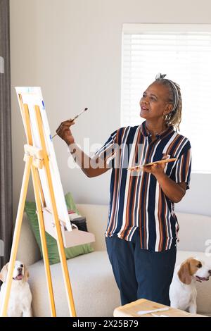 African American senior woman painting on canvas at home, dogs watching Stock Photo