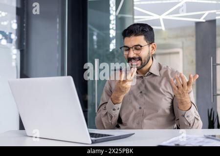 A professional man engaging with a voice assistant on his smartphone, smiling as he communicates, in a modern office setting with a laptop in front. Stock Photo