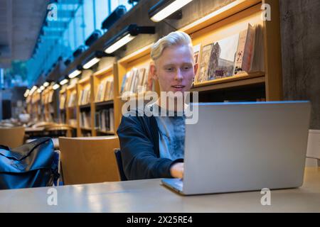 Portrait of a handsome young programmer working remotely on a netbook in a library interior, hipster guy looking at camera while updating Stock Photo