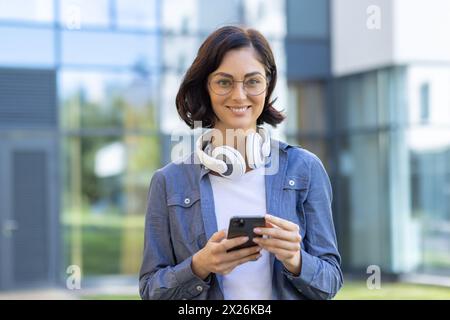 A stylish young woman in glasses and casual attire enjoys using her smartphone, with headphones around her neck, standing outdoors with a modern building background. Stock Photo