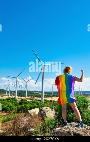 Adult woman on her back with a rainbow flag as a cape, on a rock bending her arm and showing her fist as a force in a mountainous landscape with wind Stock Photo