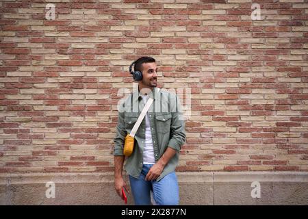 Copy space of young stylish man smiling with wireless headphones holding cell leans brick wall. Stock Photo