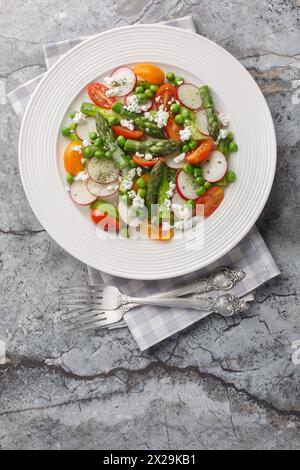 Spring salad of asparagus, radishes, cherry tomatoes, green peas and goat cheese close-up in a plate on the table. Vertical top view from above Stock Photo