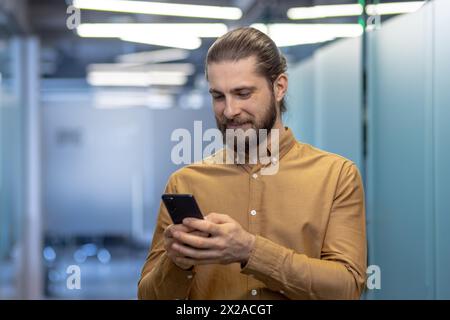A young professional man with a beard, engaged in using his smartphone, stands in a contemporary office setting. He appears focused and casual. Stock Photo