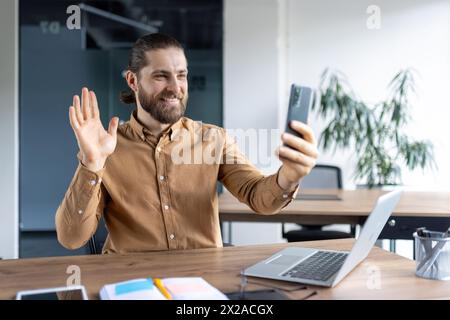 Cheerful male professional in a casual shirt waves hello during a video call on his phone, sitting at a modern workspace with a laptop and stationery. Stock Photo