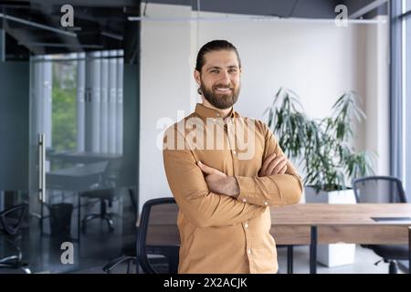 A cheerful, bearded businessman standing confidently with arms crossed in a contemporary office environment, exuding professionalism and positivity. Stock Photo