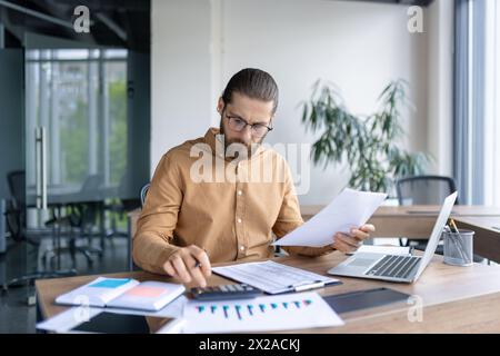 A focused young male professional examines paperwork while working in a contemporary office setting with a laptop and charts on his desk. Stock Photo