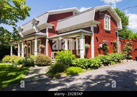 Old 1892 burgundy with white trim pinewood plank house facade with mansarde style grey sheet metal roof and landscaped front yard in summer, Quebec, C Stock Photo