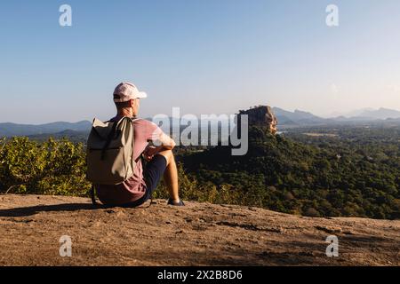 Man with backpack sitting on rock and looking at landscape. Beautiful scenery with Sigiriya rock. Solo traveler in Sri Lanka. Stock Photo