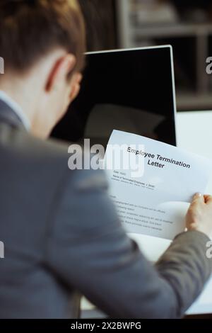 New job. Seen from behind modern 40 years old woman employee in modern green office in grey business suit with laptop and employee termination letter. Stock Photo