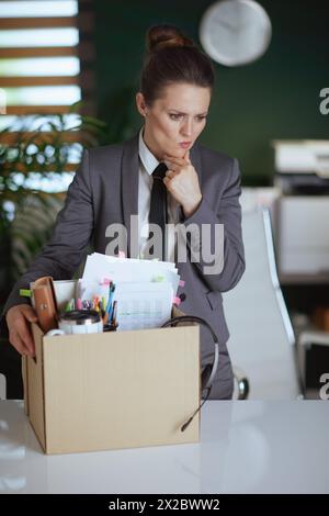 New job. pensive modern female worker in modern green office in grey business suit with personal belongings in cardboard box. Stock Photo