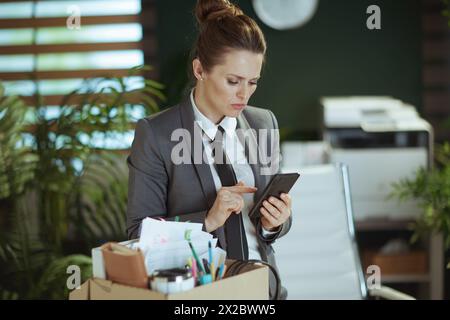 New job. pensive modern woman worker in modern green office in grey business suit with personal belongings in cardboard box using smartphone. Stock Photo