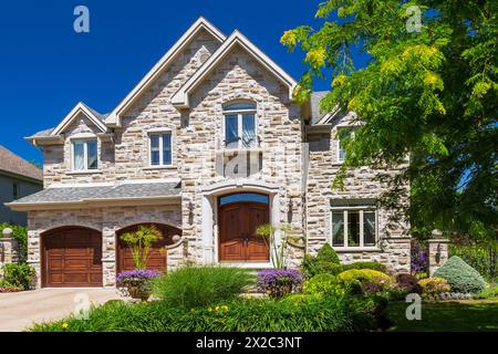 Two story beige, brown and tan cut stone with tan trim luxurious house facade with shrubs and flowers in border. Stock Photo