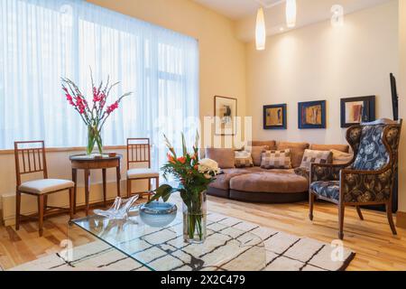 Clear glass coffee table on top of thick white rug plus Edwardian antique wooden table with chairs, upholstered sectional round sofa and arnchair. Stock Photo