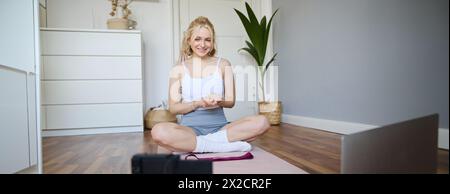Portrait of young charismatic fitness trainer, girl blogger records video on digital camera, talks about health and workout, doing exercises on rubber Stock Photo