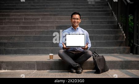 A confident Asian millennial businessman sits on outdoor steps with a coffee cup and a briefcase beside him, holding a laptop with a blank screen. bus Stock Photo