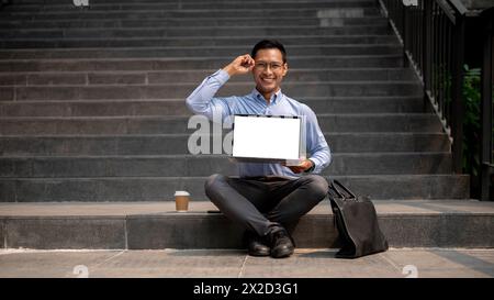 A confident Asian millennial businessman sits on outdoor steps with a coffee cup and a briefcase beside him, holding a laptop with a blank screen, and Stock Photo