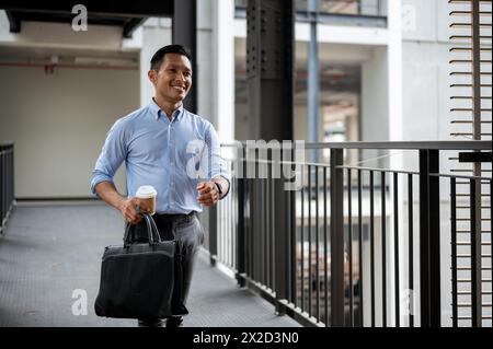 A handsome and confident Asian millennial businessman walking along the building corridor, heading to his office or leave the office, holding a coffee Stock Photo