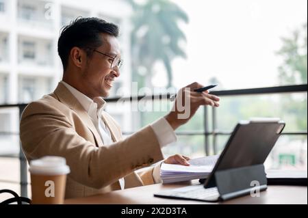 A confident, professional Asian businessman in a beige suit and eyeglasses is reviewing documents and working on a digital tablet while sitting at a t Stock Photo