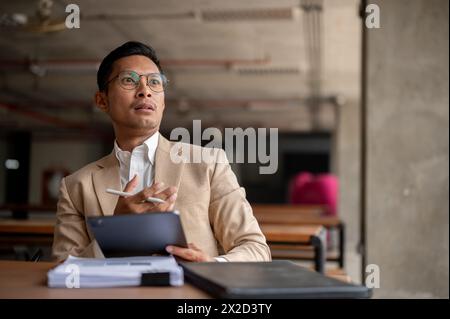 A shocked, amazed Asian millennial businessman in a beige suit and glasses looks contemplatively, holding a digital tablet and a pen, sitting at a tab Stock Photo