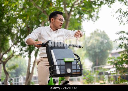 A stressed, anxious Asian millennial businessman is in a hurry to get to work in the morning, riding a bike, checking time on his wristwatch, concerne Stock Photo