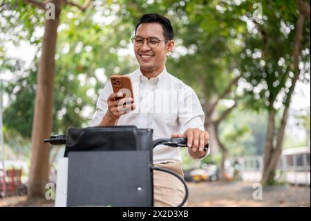 A happy, carefree Asian millennial businessman is checking messages on his smartphone while he is on a bike, riding a bike to work in the morning. mod Stock Photo