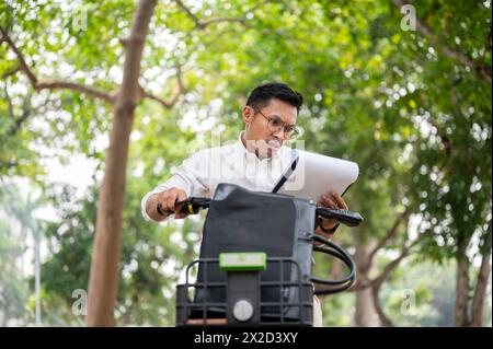 A focused, serious Asian millennial businessman rides a bike while carefully reviewing documents, multitasking, busy urgent to go to work. businesspeo Stock Photo