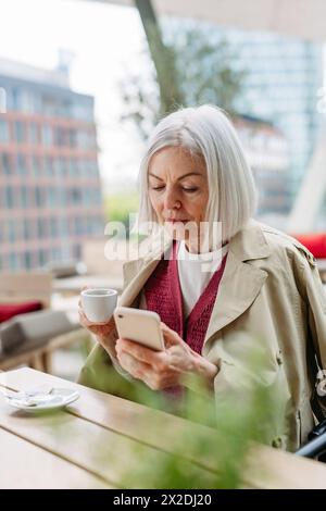 Attractive older woman checking smartphone, drinking coffee at coffee shop. Mature woman spending free time outdoors, in cafe, waiting for girl Stock Photo