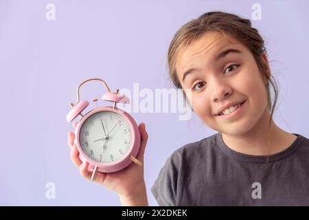 Teenage girl with desk alarm clock. Hands on the clock show 7, Close-up portrait, purple background Stock Photo