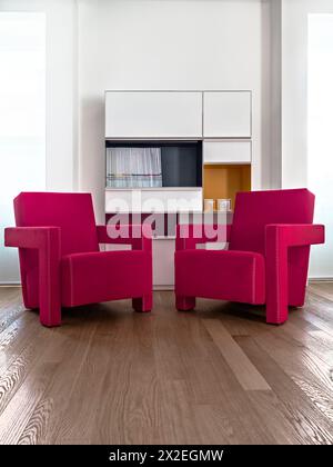 modern living room interior in the foreground two red armchair in front of a white bookcase the floor is made in wood Stock Photo