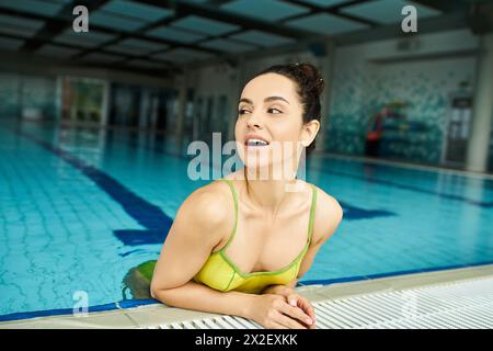 A brunette woman in a yellow bikini sitting gracefully on the pools edge in an indoor spa setting. Stock Photo