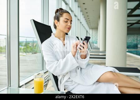 A young, brunette woman is seated in a luxurious bathrobe, engrossed in her cell phone in an indoor spa with a swimming pool. Stock Photo