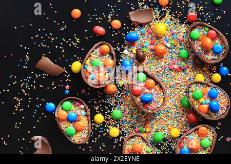 Chocolate eggs with candies, sugar colored sprinkles on a black background, top view. Easter composition. Halves of chocolate eggs on the table. Festi Stock Photo