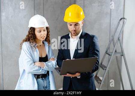 Attractive young woman contractor is talking to customer in helmet, and demonstrates sequence of repairs in living room on laptop. Meeting of landman Stock Photo