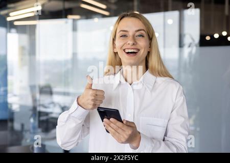 Professional young businesswoman in a white shirt giving a thumbs up while holding a smartphone. She's in a contemporary office setting with a bright smile. Stock Photo