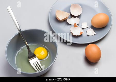 Broken egg and fork in gray bowl. Chicken shells on gray plate. Two brown eggs on table. Gray background. Top view Stock Photo