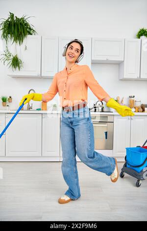 A stylish woman in casual attire gracefully mops the kitchen floor with a bucket nearby. Stock Photo