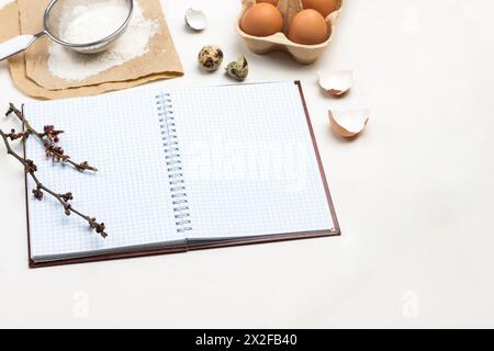 Notebook on springs. Two wooden spoons. Chicken eggs and chicken shells. Flour and sieve on paper. White background. Top view. Stock Photo