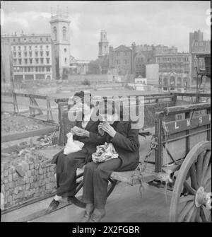 VAN GIRL: HORSE AND CART DELIVERIES FOR THE LONDON, MIDLAND AND SCOTTISH RAILWAY, LONDON, ENGLAND, 1943 - Lilian Carpenter (left) and Vera Perkins eat their packed lunch on the back of their horse-drawn LMS Railway Company van. The area behind them has been badly damaged in an air raid Stock Photo