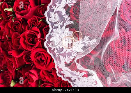 Background image for wedding design or mockup. Two gold wedding rings on a white bridal veil. Top view. red background roses bouquet. A copy space. Stock Photo