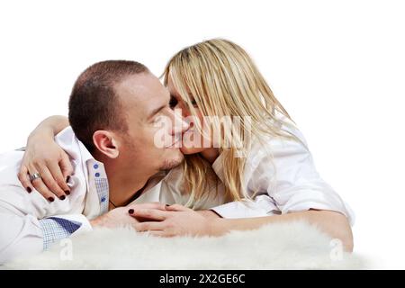 The young pair in white shirts embraces and rubs  their noses, lying on a white fleecy carpet. Stock Photo