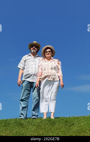 old man and woman in straw hats and sunglasses standing on summer lawn and embracing Stock Photo
