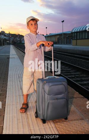 little boy stands on platform of railway with big blue travel bag Stock Photo